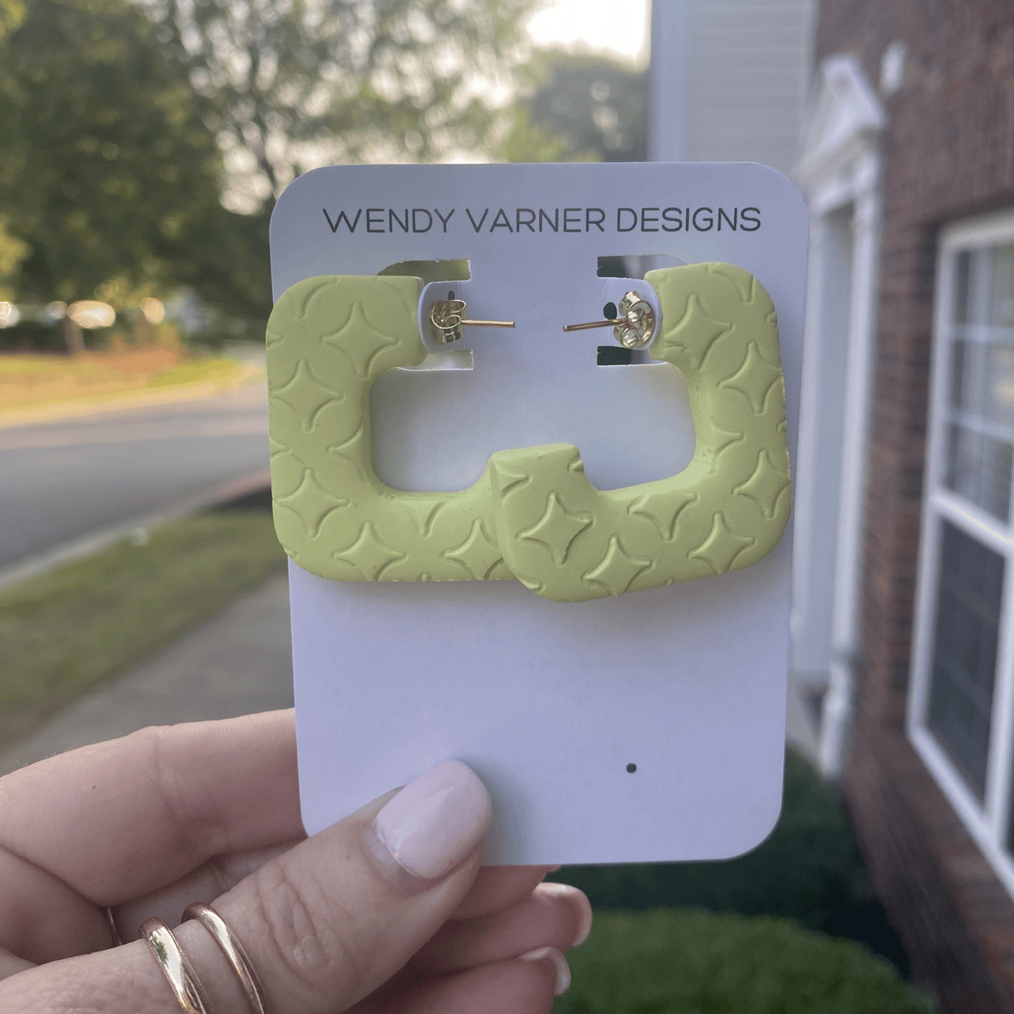 Close-up of star-textured square chartreuse earrings held against a background of greenery and neighborhood homes.