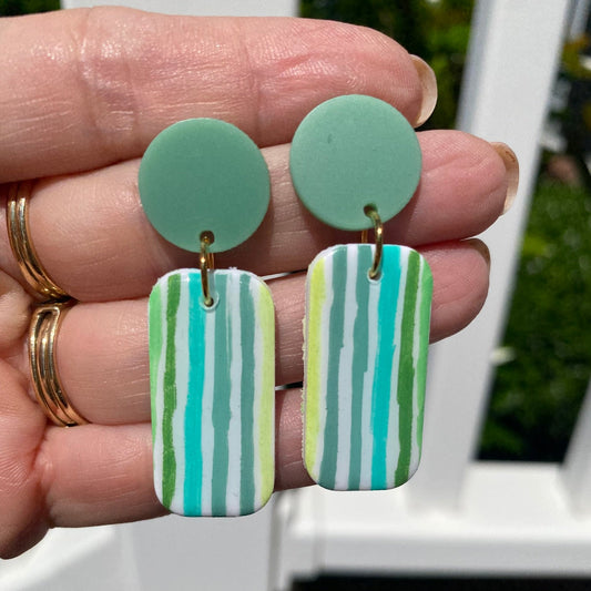 Close-up of handcrafted polymer clay stripe dangle earrings held in hand, showing mint, teal, olive, and yellow-green stripe detail