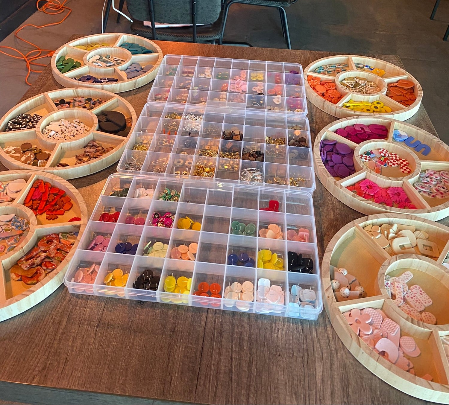 Table with craft supplies including colorful polymer clay earrings and a clear organizer in a room with people sitting at tables.