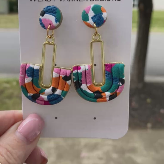 Close-up of handmade polymer clay earrings featuring pink, teal, orange, black, and blue pattern