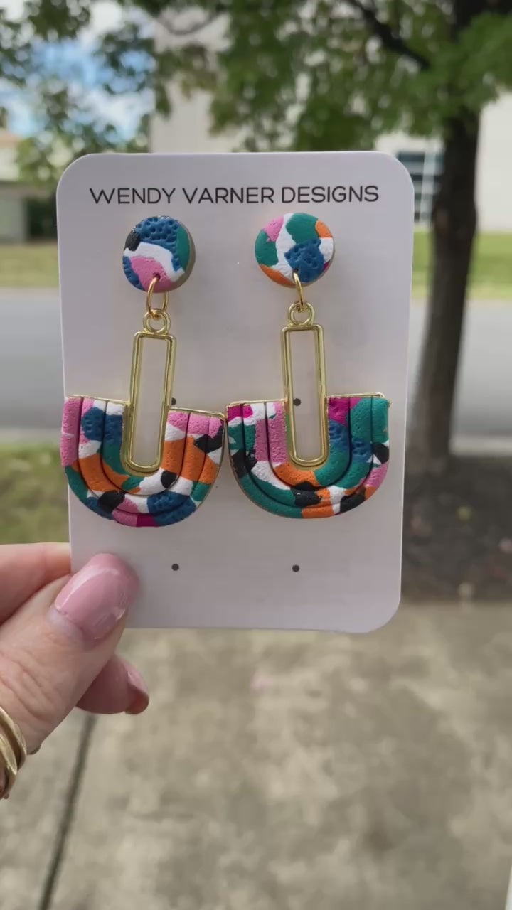 Close-up of handmade polymer clay earrings featuring pink, teal, orange, black, and blue pattern